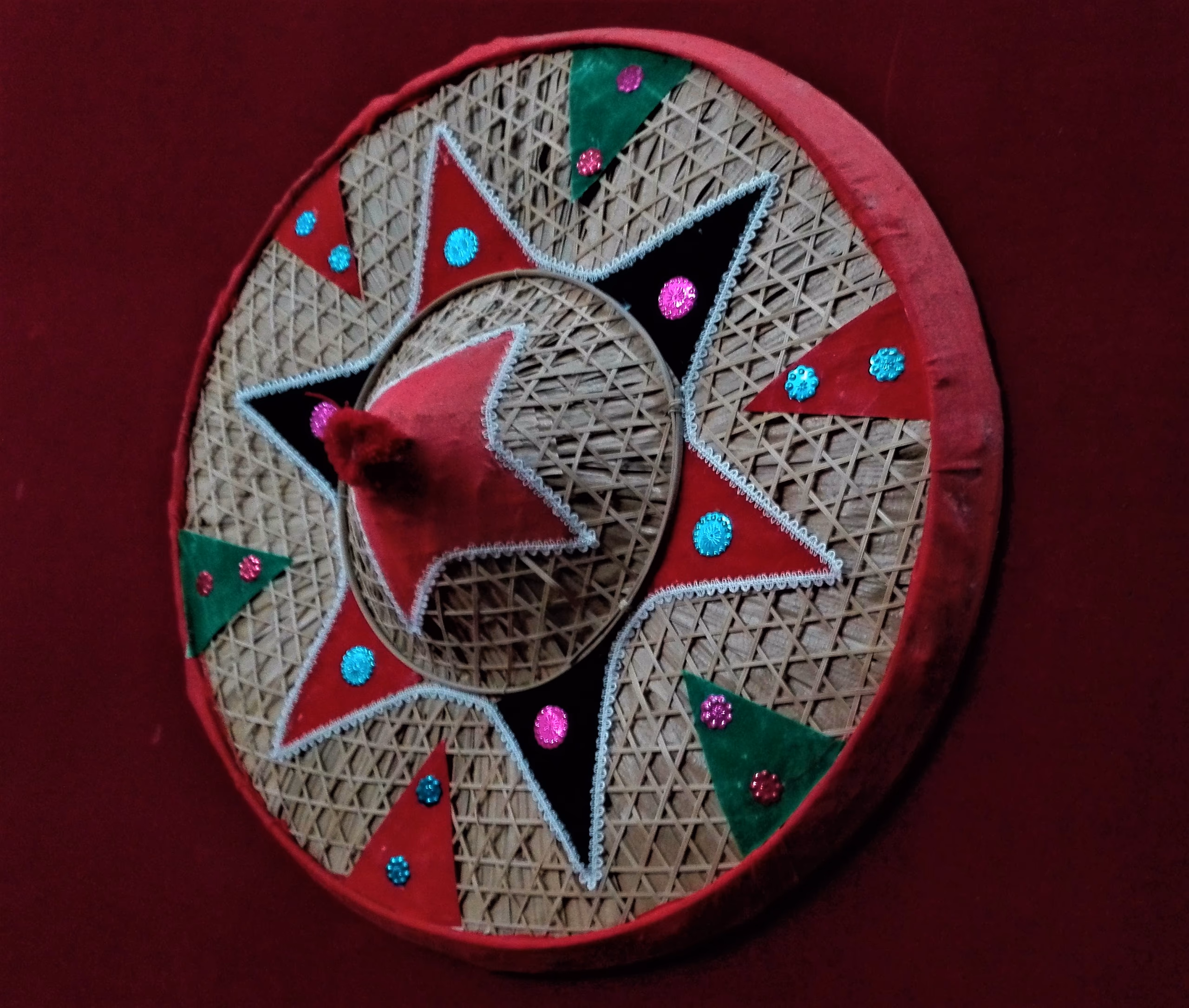 A round phulam japi faced in woven bamboo with a red rim, a green-and-red appliqué star, small mirrors and sequins, set against a deep maroon backdrop