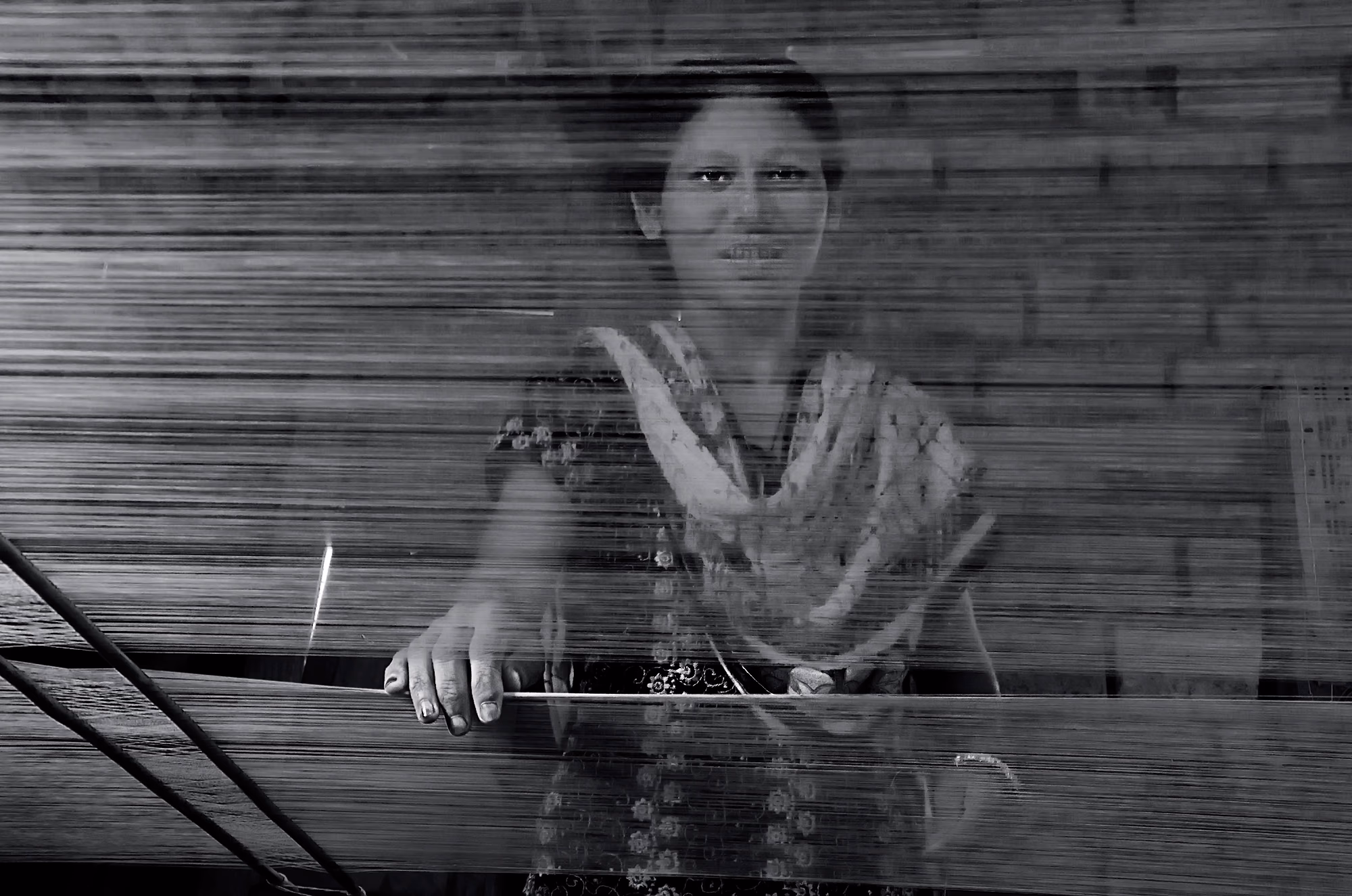 A weaver seen through the stretched warp threads of a handloom, her hand resting on the loom beam, in Sualkuchi