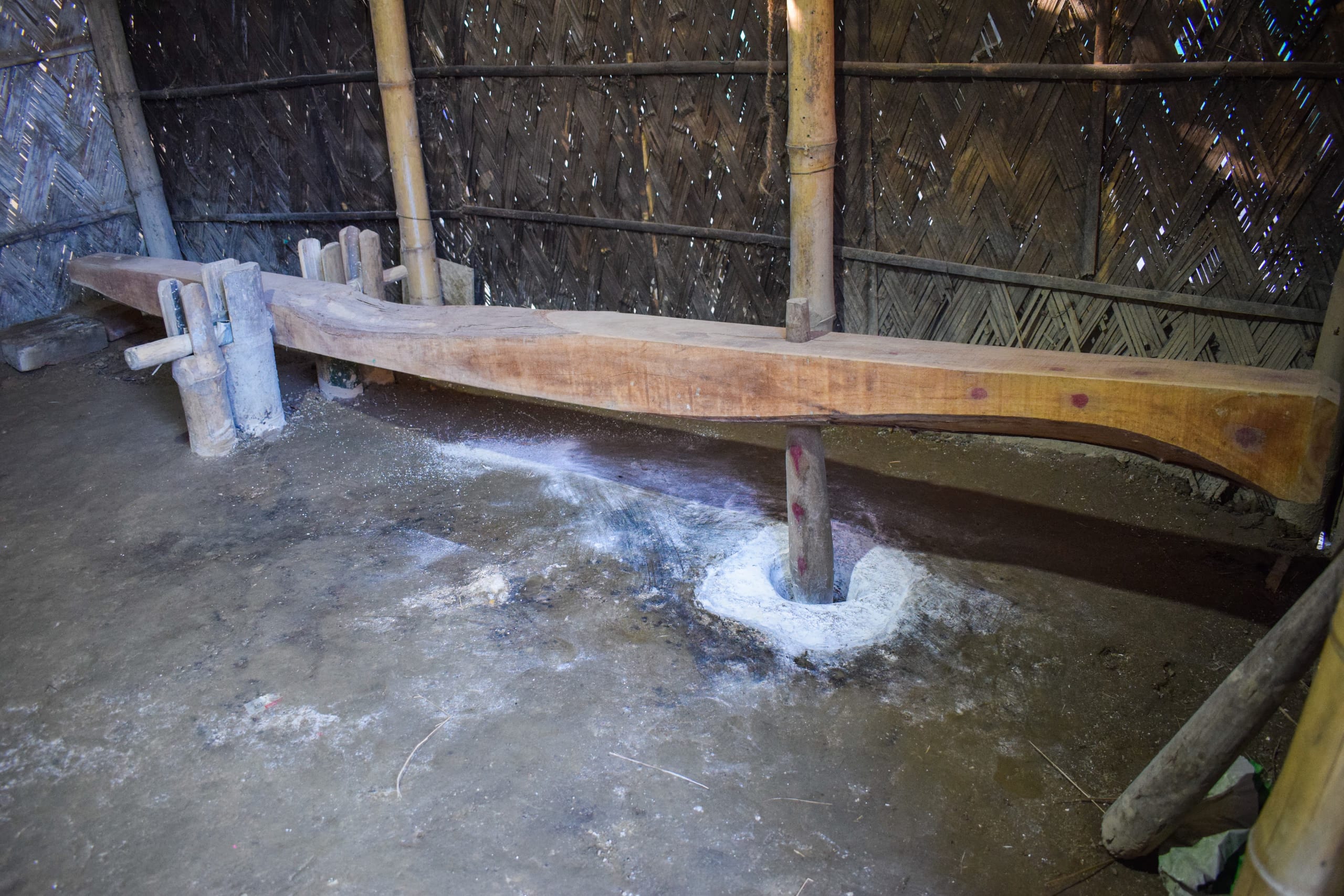 A wooden dheki, a foot-operated lever pestle, set over a floor mortar inside a bamboo-walled house