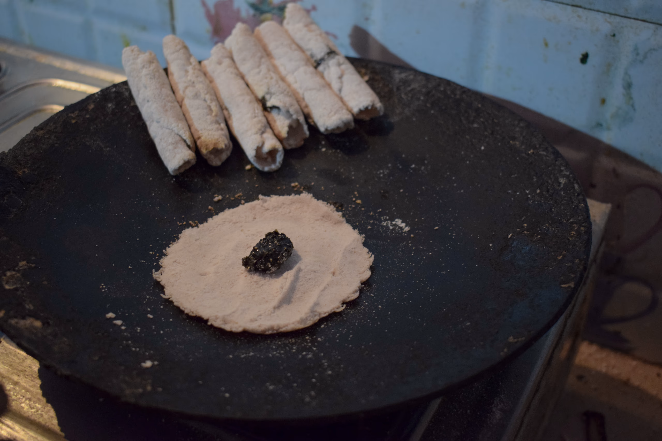 Rolled til pitha at the back of a hot griddle and, in front, an open round of rice-flour batter with a dollop of dark sesame-and-jaggery filling ready to be folded