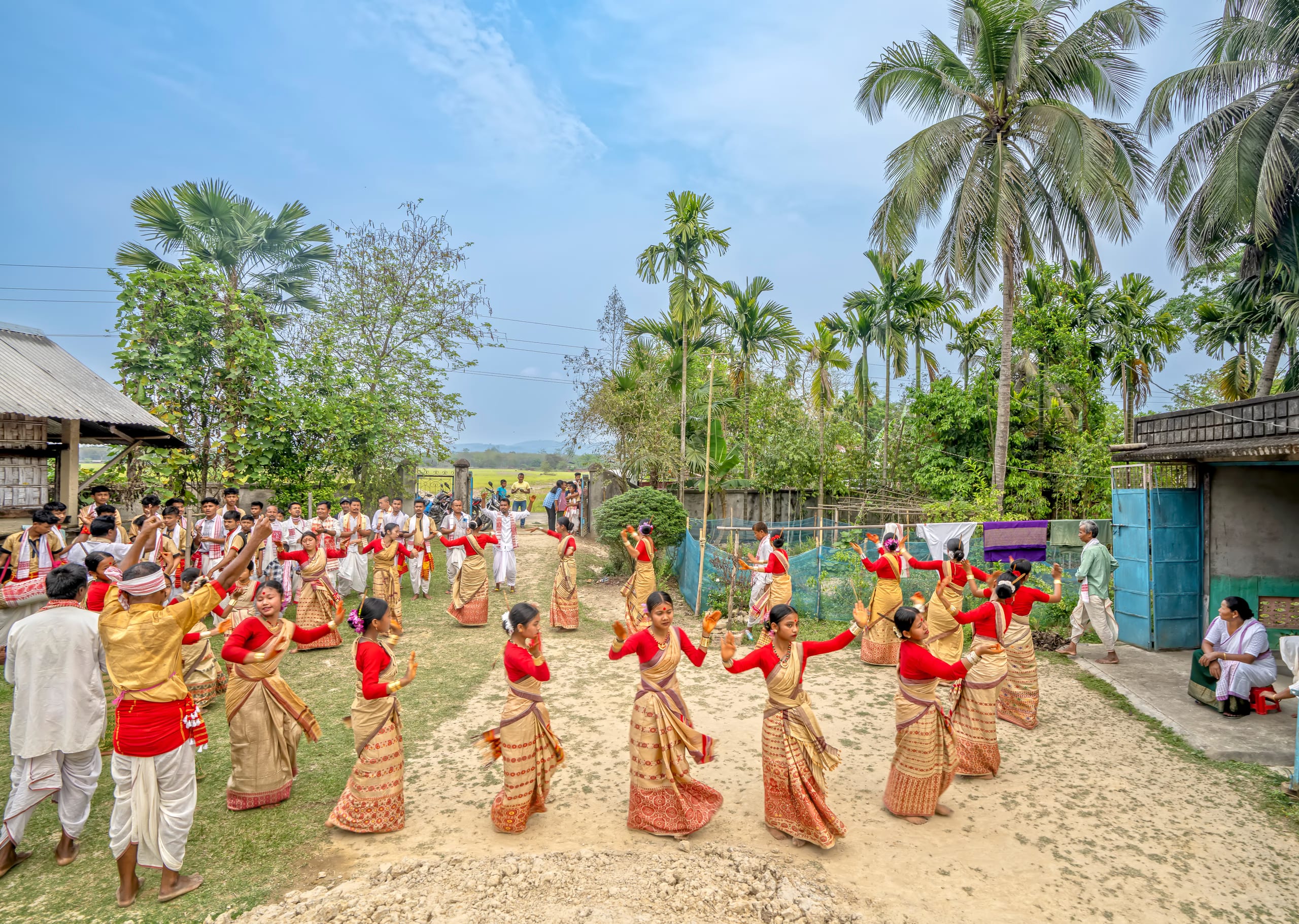 Young women in red-and-cream mekhela sador dancing in a ring in a village yard, with male drummers at the edge
