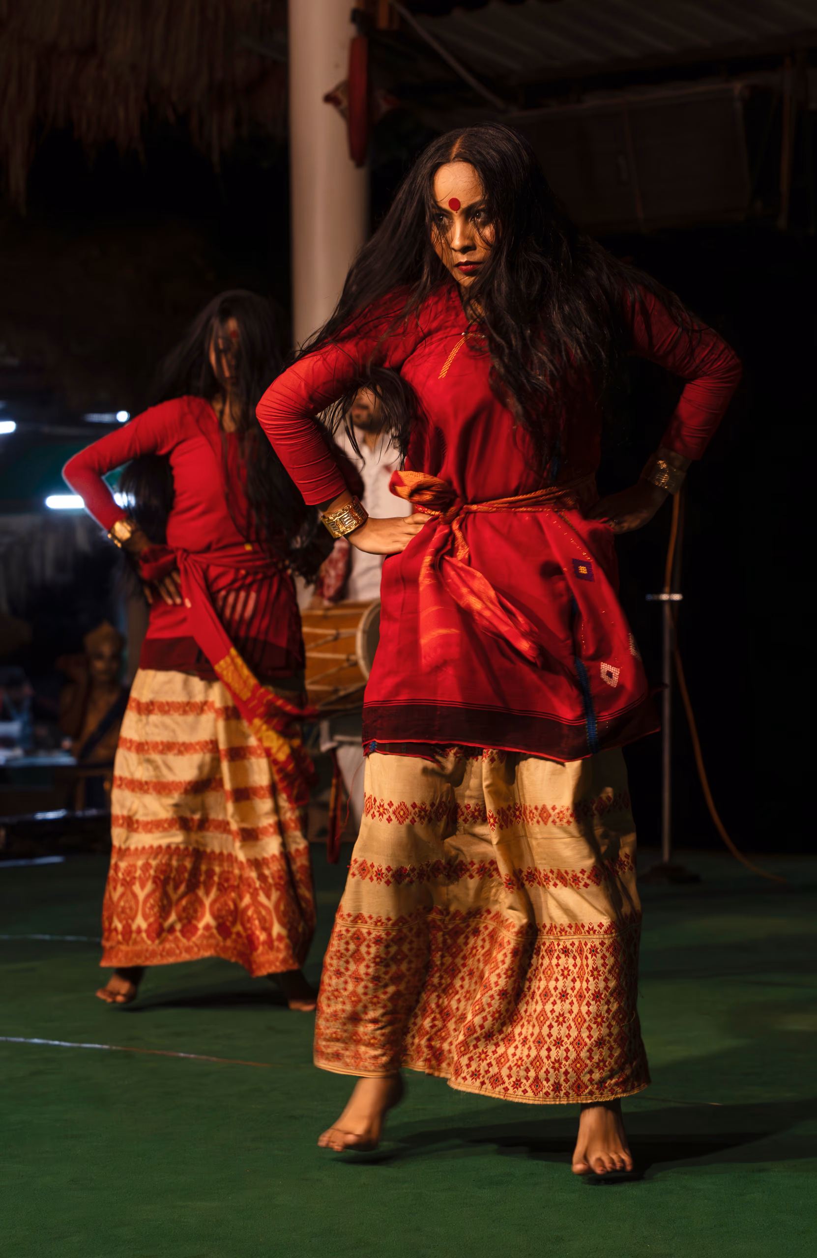 A Sattriya dancer in a red blouse and cream Muga silk skirt woven with traditional Assamese motifs, gold cuff bracelets, a khol drum visible behind