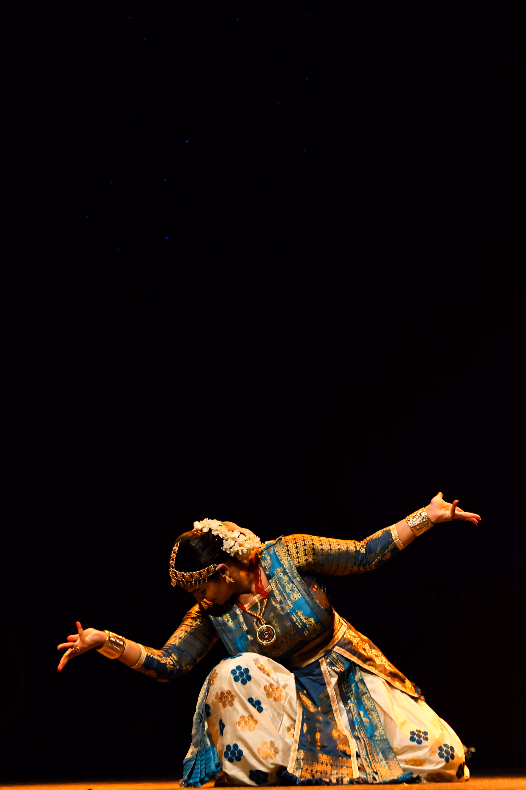 A Sattriya dancer in a blue and gold silk costume with a white floral Muga skirt, kneeling low with both arms extended in a graceful pose against a dark stage