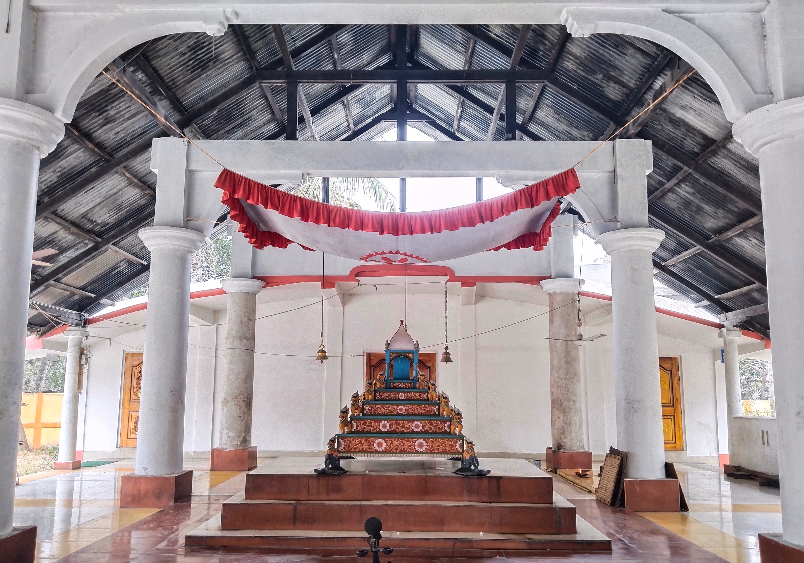 Interior of a Vaishnavite namghar prayer hall with white pillars and a central decorated altar throne flanked by hanging bells