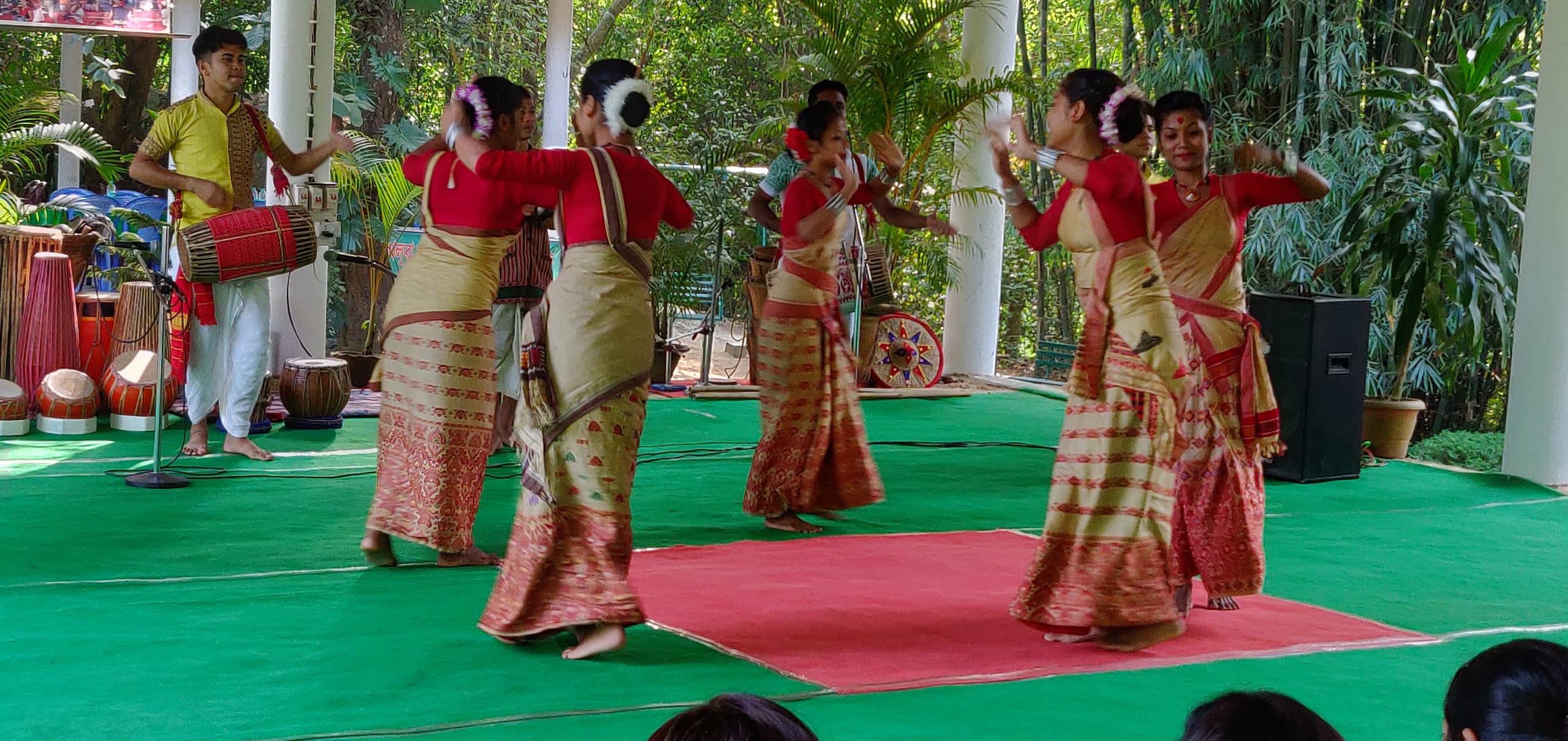 Women in red-and-cream mekhela-chador dancing Bihu on a green-carpeted stage while a man plays the dhol drum at left