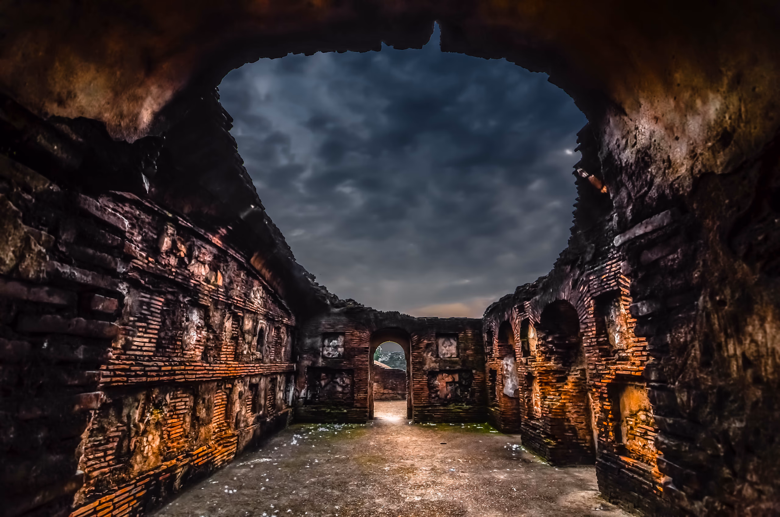 Vaulted brick chamber of the Talatal Ghar opening to a stormy sky through a broken roof, warm-lit golden brick walls and a doorway of light at the far end