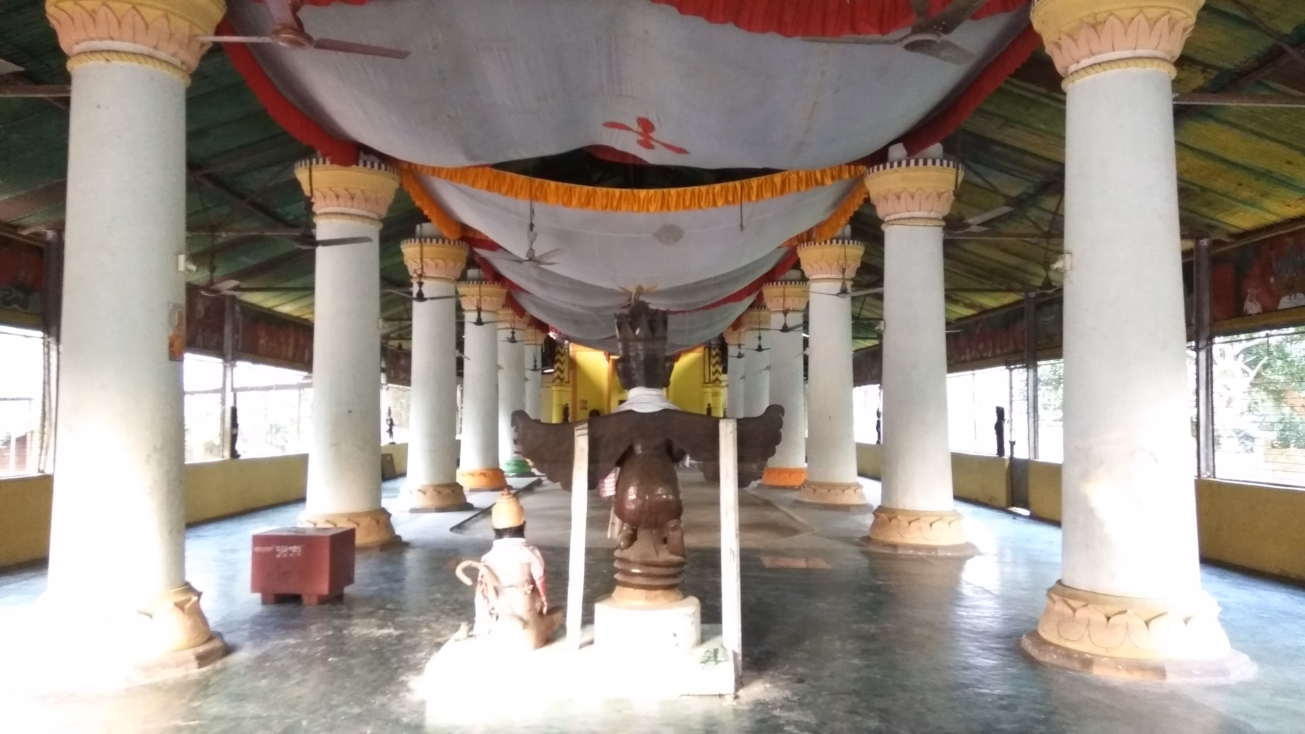 The pillared interior of a Majuli sattra namghar, two rows of whitewashed columns with painted lotus capitals under a draped red-and-white ceiling canopy, a carved wooden throne-pedestal standing in the central aisle on a polished floor