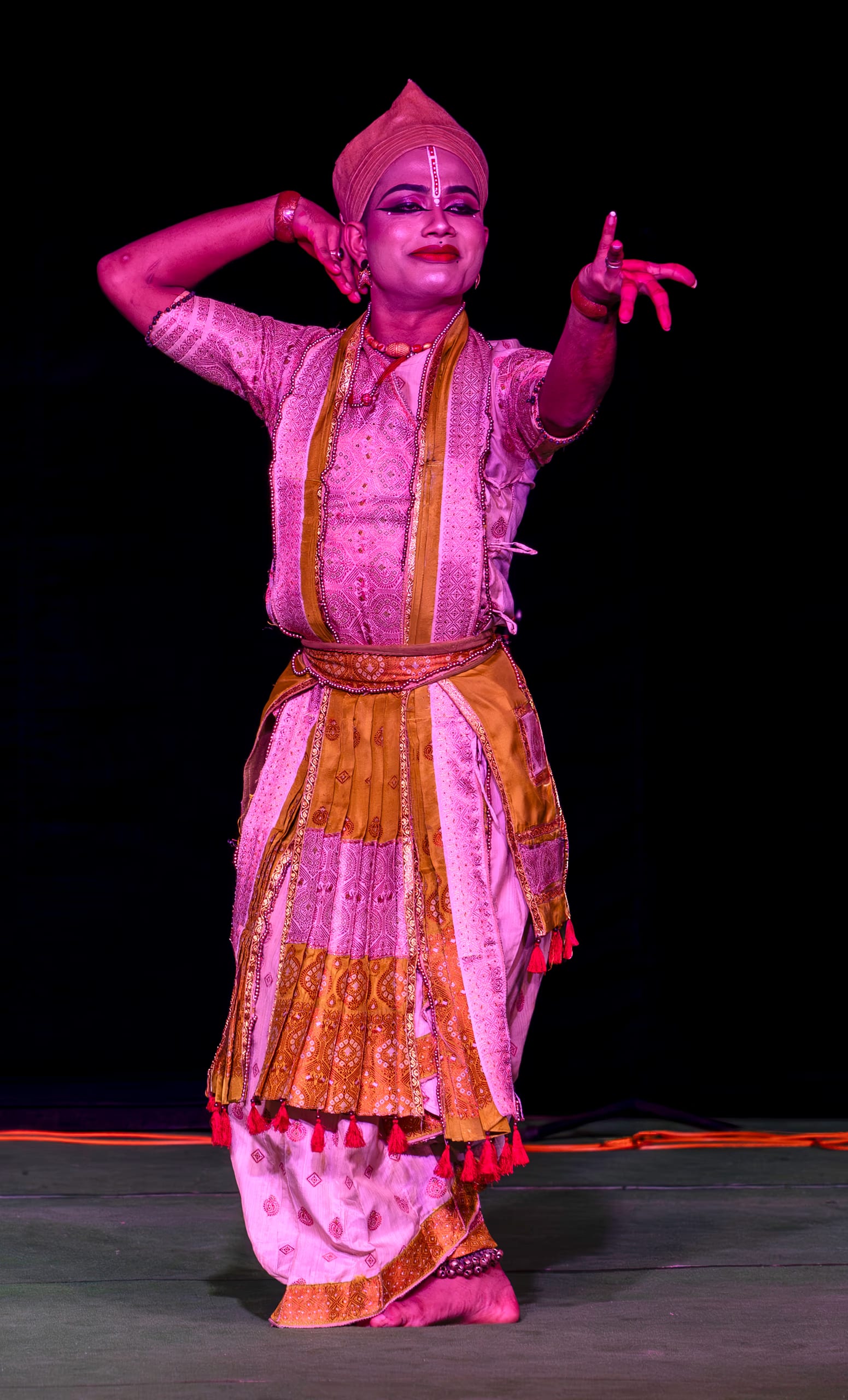 A solo Sattriya dancer in a conical cloth turban and pleated gold-and-cream silk costume, one arm raised overhead and the other extended in a mudra, performing on a dark stage