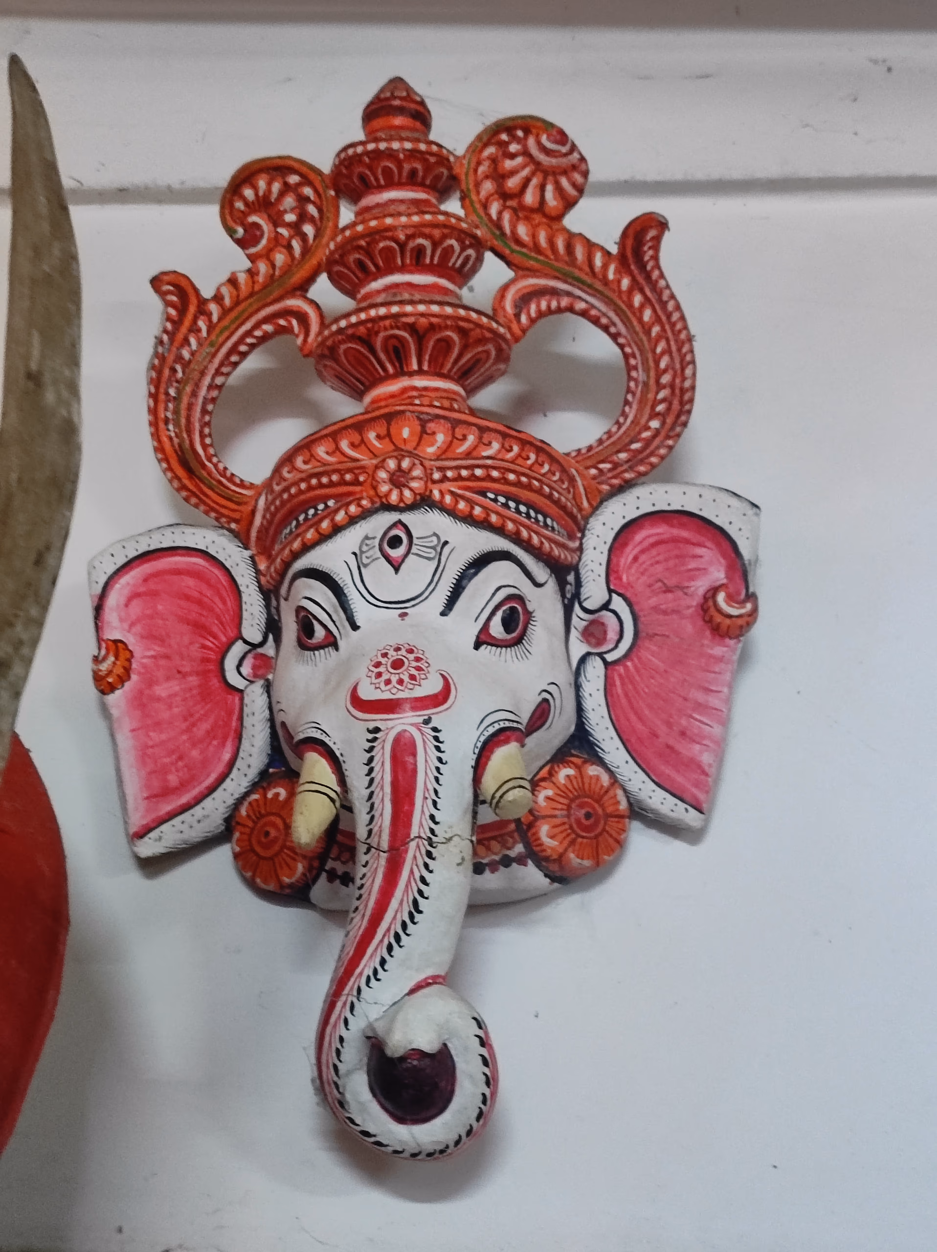 Painted Ganesha mukha: a pale elephant face with a long curling trunk, pink fanned ears, almond eyes, and a tall ornate red-and-orange crown, set against a white wall