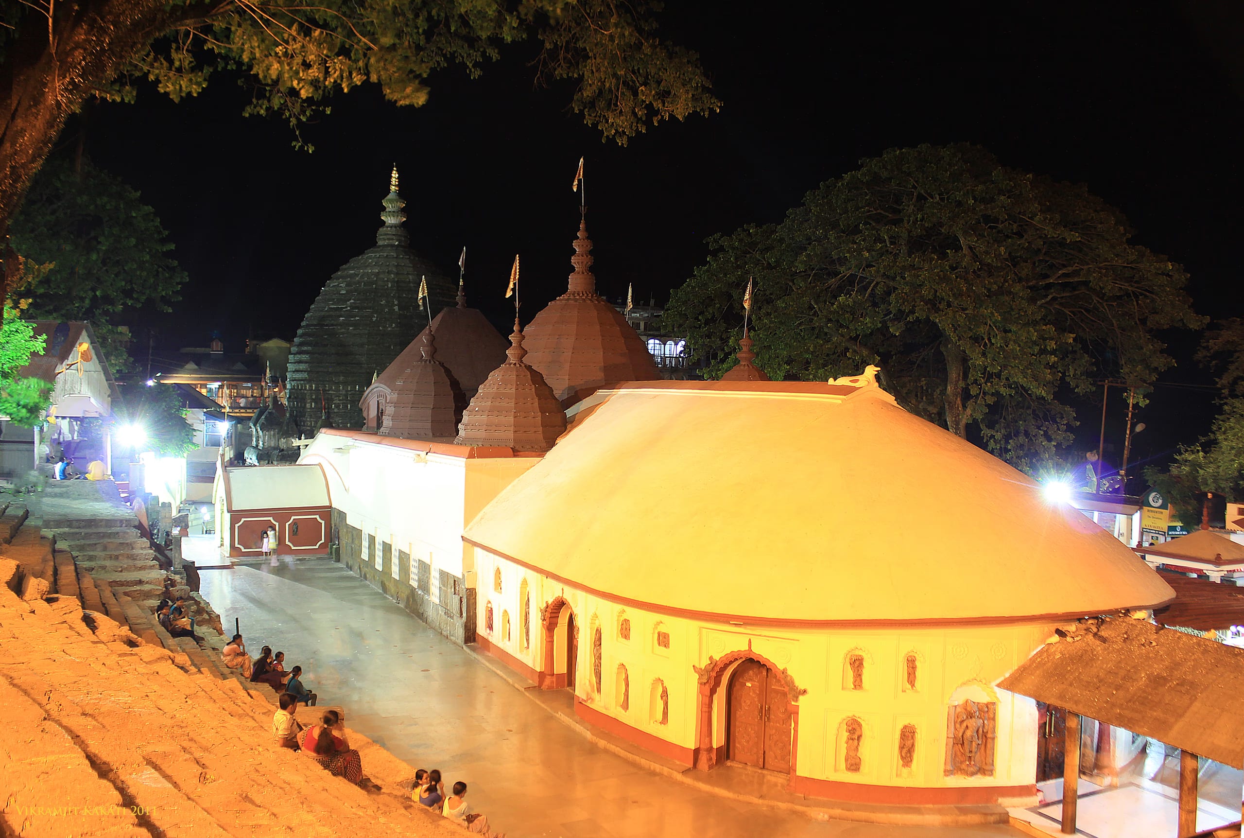 The Kamakhya temple complex lit at night: a cream-and-gold mandapa with a red-banded base in front, dark stone beehive shikharas of the sanctum and subsidiary shrines rising behind, stone steps descending at left