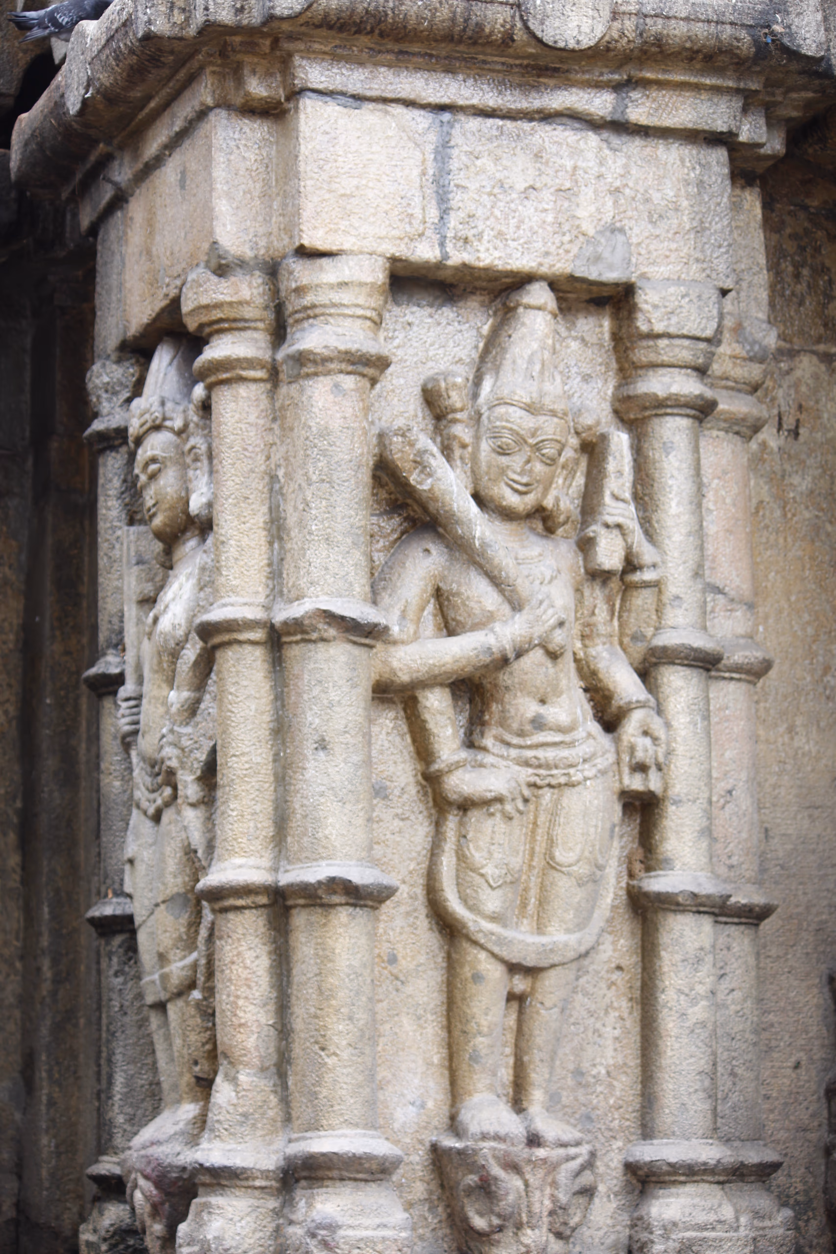 A weathered grey stone relief of a standing crowned deity figure carved on the exterior wall of the Kamakhya temple, set between two slender carved pilasters, one arm raised